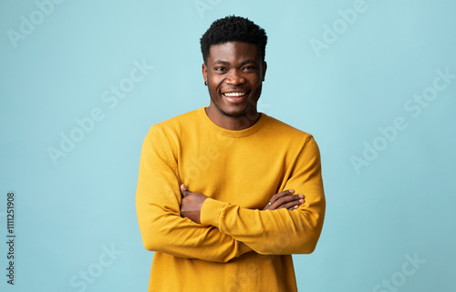 Cheerful handsome young african american guy in yellow sweater posing alone on blue studio background, holding hands crossed on chest and smiling at camera, copy space for advertisement