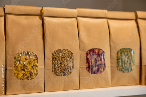 A row of brown paper bags with transparent windows reveals various dried herbs and flowers in yellow, purple, green, and orange hues, neatly arranged on a shelf. photo