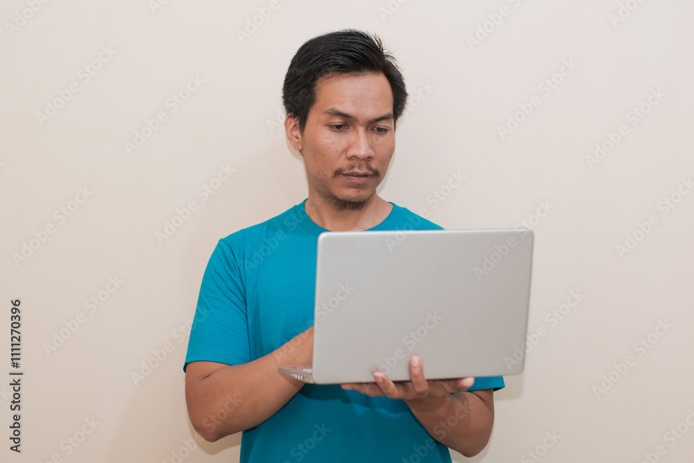 Smiling Southeast Asian man wearing green t-shirt casual clothes holding laptop, standing over isolated white background, Positive person Smiling Southeast Asian man wearing green t-shirt casual clothes holding laptop, standing over isolated white background, Positive person