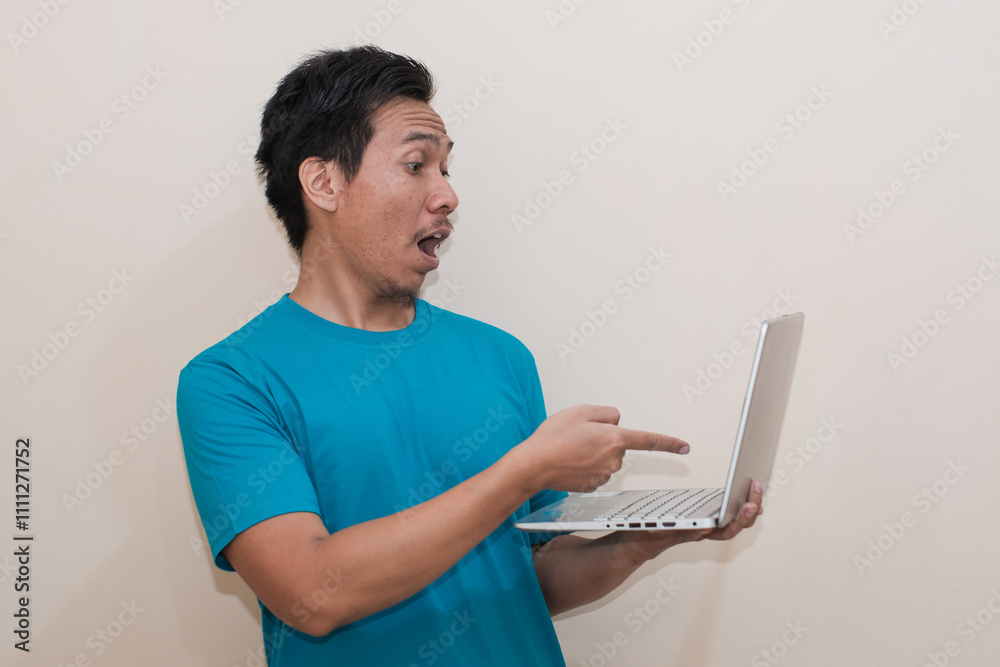Shocked Southeast Asian man wearing green t-shirt casual clothes holding laptop and pointing at laptop, standing over isolated white background, Positive person Shocked Southeast Asian man wearing green t-shirt casual clothes holding laptop and pointing at laptop, standing over isolated white background, Positive person