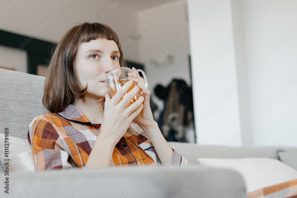 Young Woman Feeling Unwell, Holding A Cup Of Tea And Suffering From A Headache While Resting On A Sofa At Home Young Woman Feeling Unwell, Holding A Cup Of Tea And Suffering From A Headache While Resting On A Sofa At Home