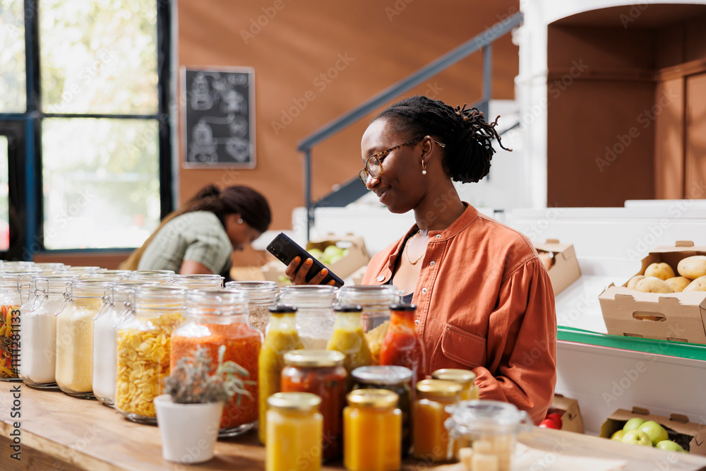 Young black woman using smartphone while browsing through the eco friendly store. African american female customer surfing the net on mobile device for healthy bio food recipes. Young black woman using smartphone while browsing through the eco friendly store. African american female customer surfing the net on mobile device for healthy bio food recipes.
