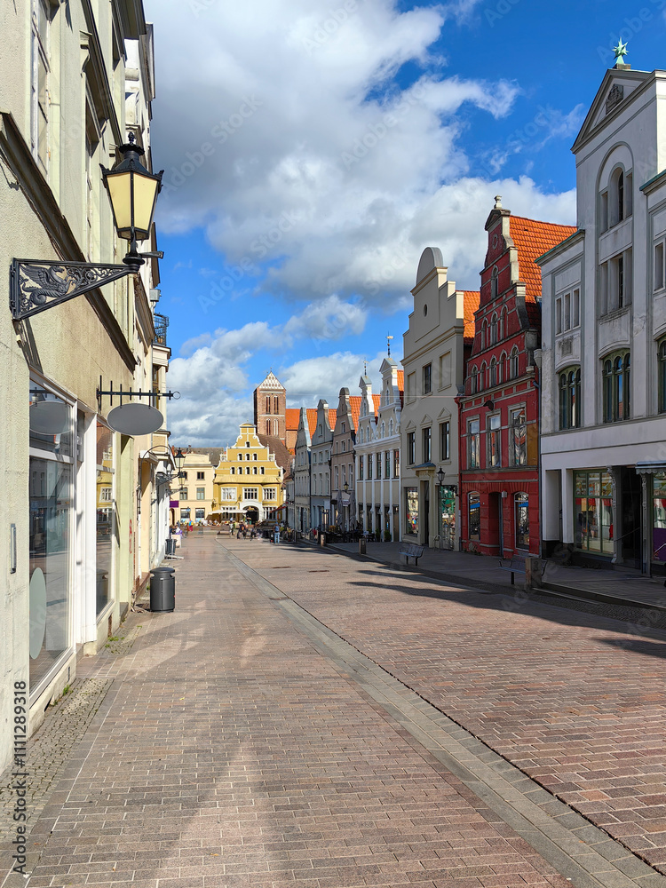 Obraz premium A picturesque view of Krämerstraße in Wismar, Germany, showcasing historic architecture, cobblestone streets, and vibrant storefronts under a clear sky. Perfect for travel and cultural themes.