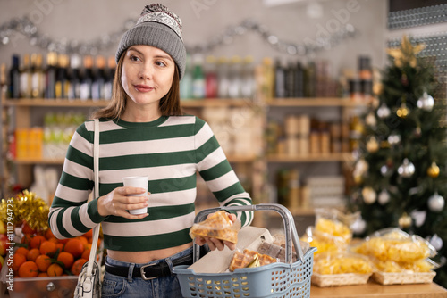 While shopping before new year, girl in winter hat walks through grocery store, studies windows, looks for something in store, drink hot coffee, spending time in moll shopping center.