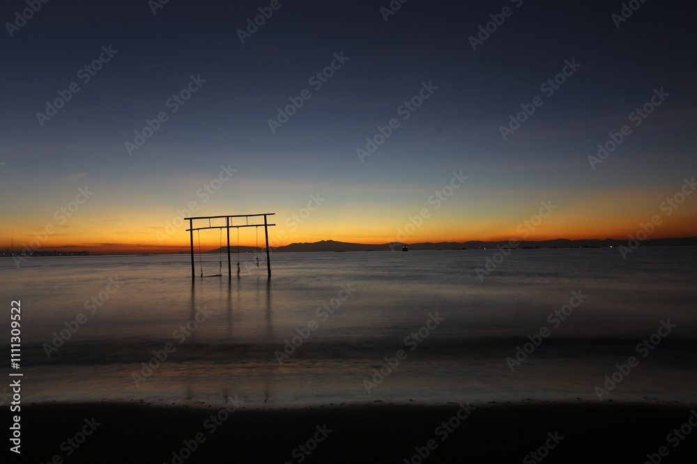 A tranquil sunrise over a still lake features the silhouette of an abandoned swing set.