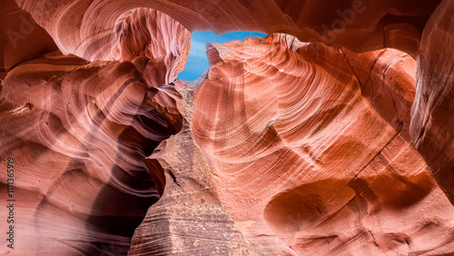 This Antelope slot canyon near Page, AZ shows flowing sandstone walls in warm hues, lit by sunlight, contrasts with a bright blue sky peeking through narrow gaps.