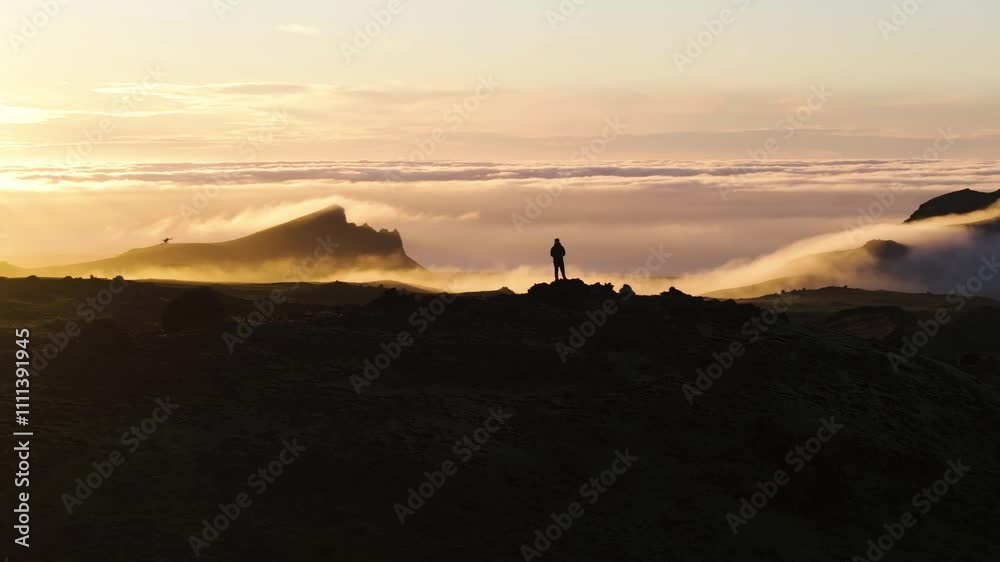 AERIAL: Person standing on top of a mountain above clouds, sunrise in Iceland AERIAL: Person standing on top of a mountain above clouds, sunrise in Iceland