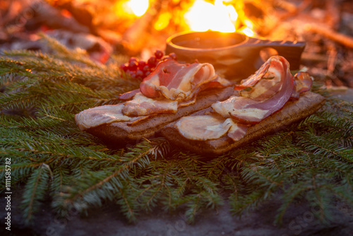 Food in the forest. Bacon strips on a piece of bread on spruce branches against the backdrop of a fire. Kuksa of forest tea