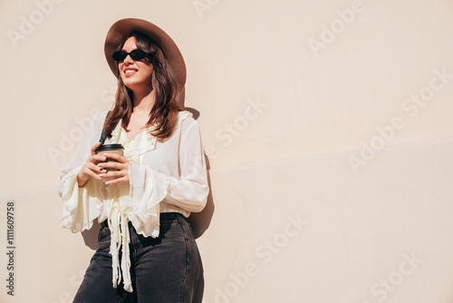 Lifestyle portrait of young stylish woman dressed in white blouse. Model walking with coffee cup in old town in sunny day. Beautiful smiling female in hat. Cheerful and happy. Takeaway coffee, eyewear