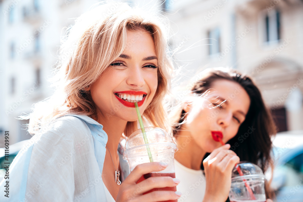 Two young beautiful smiling glam female in trendy summer clothes. Sexy women posing outdoors. Positive models holding and drinking fresh cocktail smoothie drink in plastic cup. Bright makeup, red lips Two young beautiful smiling glam female in trendy summer clothes. Sexy women posing outdoors. Positive models holding and drinking fresh cocktail smoothie drink in plastic cup. Bright makeup, red lips