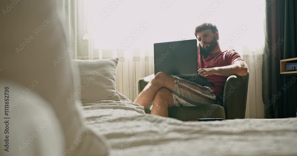 Young man working from home. He's sitting in his bedroom, wearing casual clothes and using a laptop. The room is bright and airy. Young man working from home. He's sitting in his bedroom, wearing casual clothes and using a laptop. The room is bright and airy.