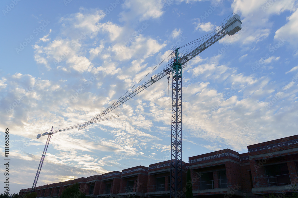 silhouette of two construction cranes against a blue sky with clouds silhouette of two construction cranes against a blue sky with clouds