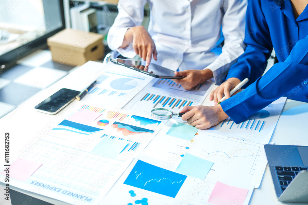 Two Asian business woman working and talking at a table with documents, a graph, and a digital tablet. They are focused on analysis, business planning, and collaborative discussions Two Asian business woman working and talking at a table with documents, a graph, and a digital tablet. They are focused on analysis, business planning, and collaborative discussions
