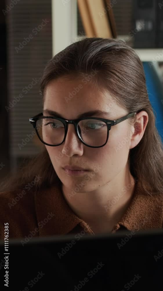 Vertical chest up of focused young Caucasian woman wearing glasses and brown polo sweater working at dual monitors in modern office environment
