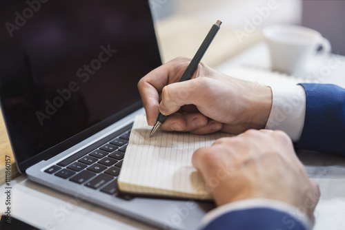 The image focuses on a man's hand scribbling in a notepad on a stylish laptop, set against a blurry scene photo