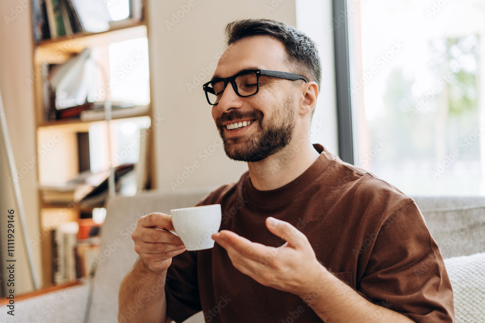 Smiling Latin man wearing eyeglasses sitting at table, closing his eyes and holding cup of coffee Smiling Latin man wearing eyeglasses sitting at table, closing his eyes and holding cup of coffee