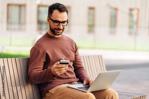 Smiling man, wearing stylish casual clothes and eyeglasses, using laptop, holding mobile phone