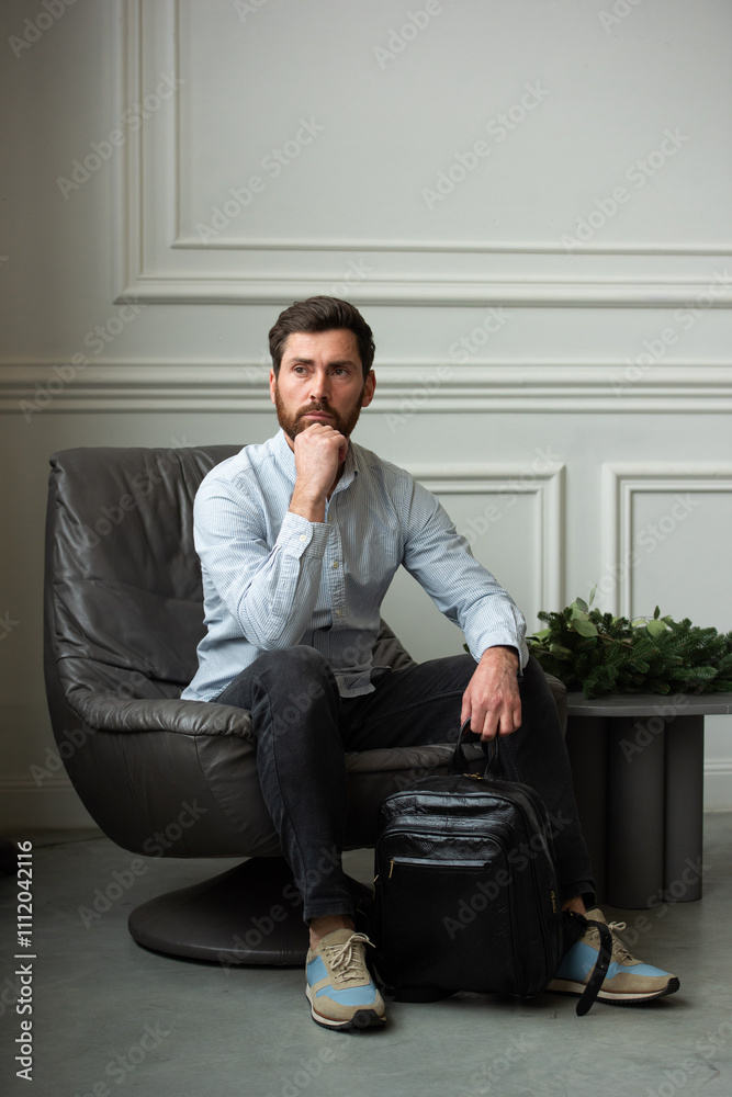 Fototapeta premium A man is comfortably sitting in a chair, resting with a leather backpack near him