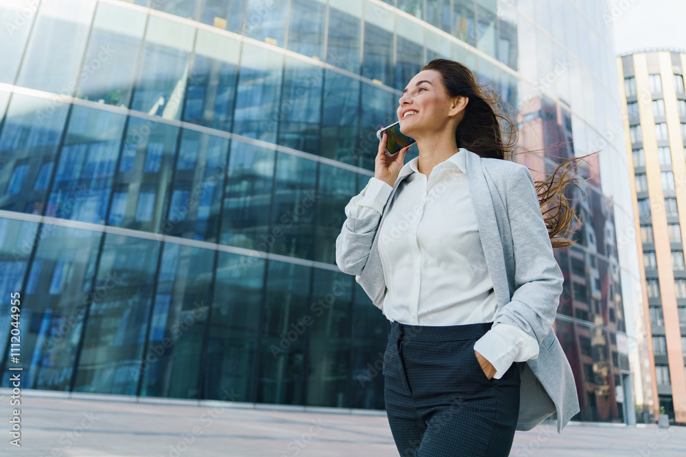 Businesswoman walks confidently while talking on her smartphone near modern glass buildings in an urban setting Businesswoman walks confidently while talking on her smartphone near modern glass buildings in an urban setting