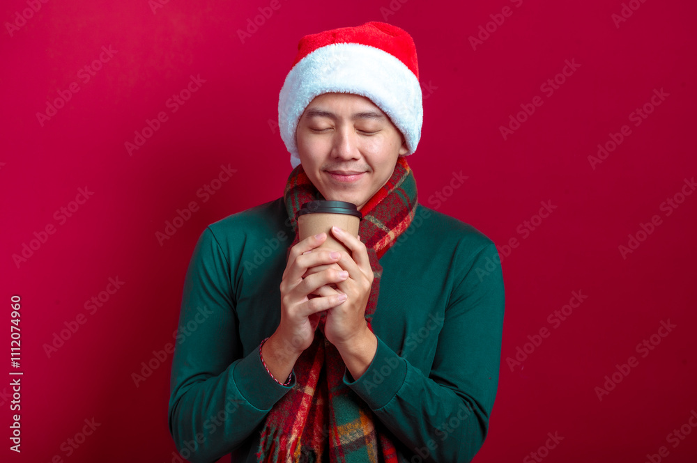 An Asian man in a Santa hat, green sweater, and plaid scarf smiling warmly while holding a coffee cup, with a red background conveying a festive and cozy Christmas vibe