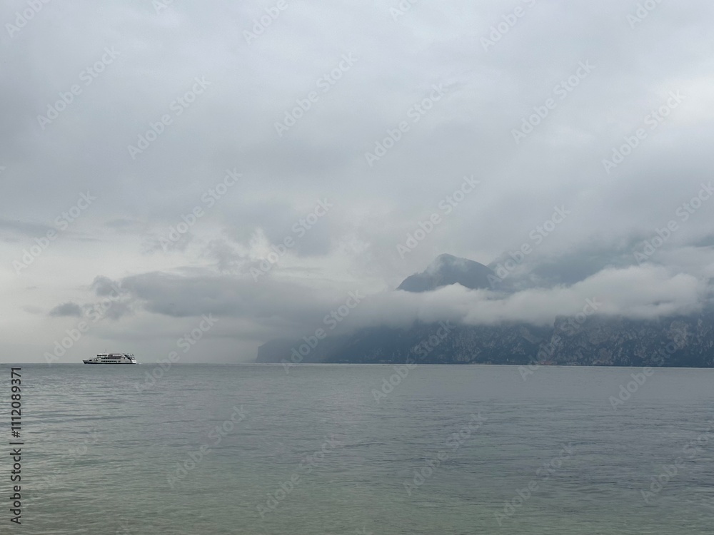 A boat is sailing on the Lake Garda under the grey clouds over the surrounding mountains, Malcesine, Veneto, Italy, October 2023