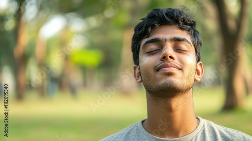 Close-up photo of a happy and satisfied young Indian man standing in a park with his eyes closed, resting and breathing deeply