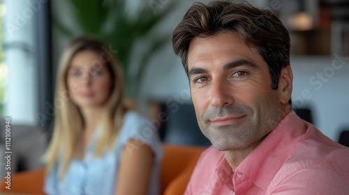 Confident man in a relaxed setting with a woman out of focus in the background.