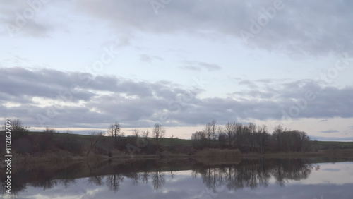 A tranquil scene featuring a calm lake reflecting bare trees and a cloudy sky in the evening before twilight. The horizon shows rolling hills, creating a peaceful and picturesque natural setting.