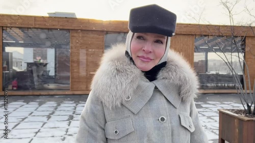 Portrait trendy mature woman Stands outside in snow, wears fashionable winter multilayer outfit, grey coat with fur collar, hood and stylish black cap. Female warm clothes