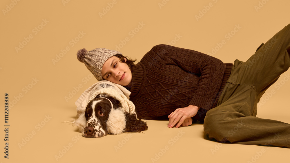 Young man in warm chocolate sweater and hat, posing with adorable calm dog wearing sweater and warm headwear, against beige studio background. Concept of animal, winter season, friendship, happiness Young man in warm chocolate sweater and hat, posing with adorable calm dog wearing sweater and warm headwear, against beige studio background. Concept of animal, winter season, friendship, happiness