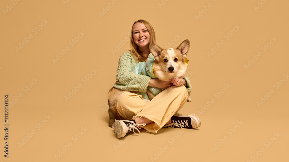 Portrait of happy smiling young woman in light green cozy cardigan, sitting on floor and posing with adorable Corgi dog against studio background. Animal, winter season, friendship, happiness concept Portrait of happy smiling young woman in light green cozy cardigan, sitting on floor and posing with adorable Corgi dog against studio background. Animal, winter season, friendship, happiness concept