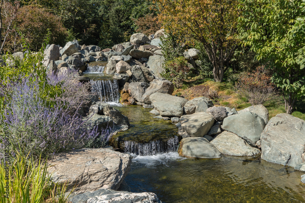 Water falls from height of 7 meters into artificial rocky riverbed. Close-up. Triple waterfall in Japanese Garden. Riverbed is divided into three streams, which turn into waterfalls. Water falls from height of 7 meters into artificial rocky riverbed. Close-up. Triple waterfall in Japanese Garden. Riverbed is divided into three streams, which turn into waterfalls.