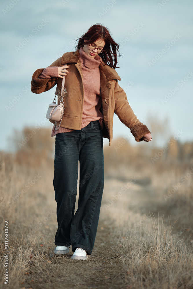 Teenage girl with red hair and glasses walking along a rural path, holding her brown coat while carrying a small bag, with her hair blowing in the wind on an autumn day.