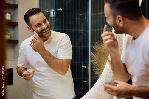 Fotografia Reflection in  mirror of happy man applying face cream in  morning
