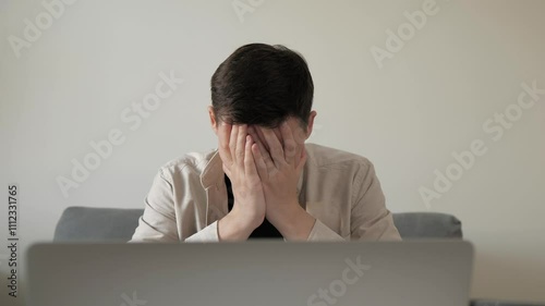 Young man feeling stressed while working on laptop in a modern home interior. Disappointed man covering face palm thinking. Depressed businessman looking frustrated.