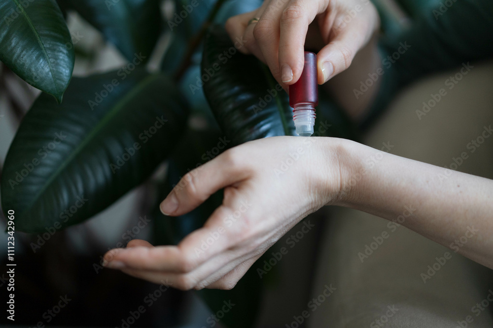 Obraz premium A gentle hand rolls essential oil onto the wrist in a serene indoor environment. Lush green leaves provide a calming backdrop, enhancing the sense of relaxation and wellness.