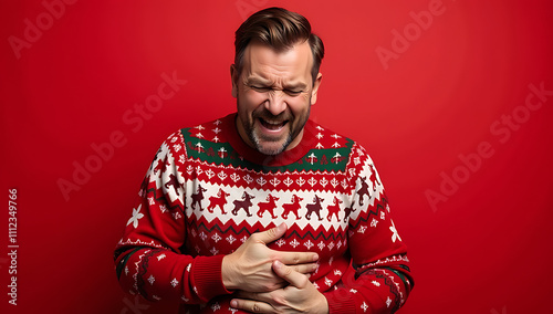 Man in Christmas sweater clutches his hurt stomach on a bright red background, eyes scrunched up in apparent discomfort._00001_