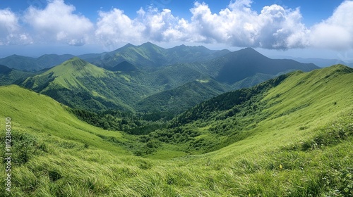 A panoramic view of rolling green hills and mountains under a bright blue sky with puffy white clouds.