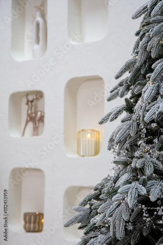 Modern Christmas interior with a focus on details: snow-covered branches of a Christmas tree with shimmering garlands and pearl decor. In the background, a wall with decorative niches decorated with a