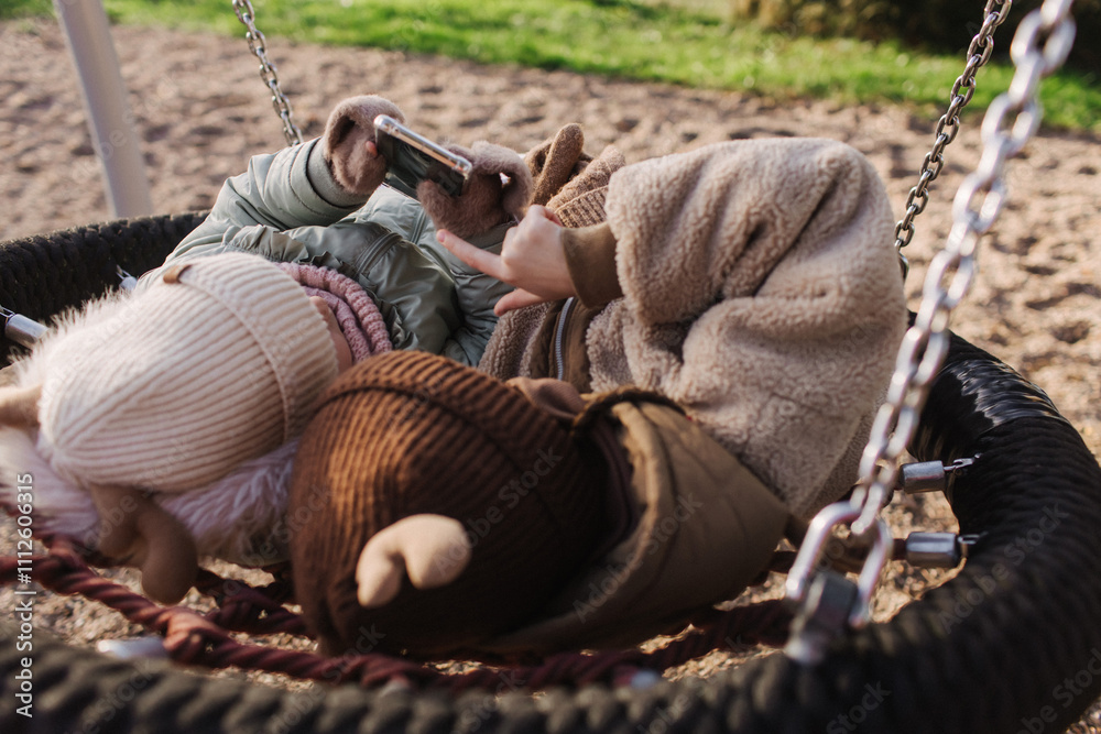 Fototapeta premium Cute little girls, two preschooler female kids uses the phone and headphone in the school yard. Concept image for child development, healthy kid playing outside. Gen Alpha. Part of a series