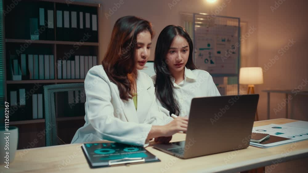 Two businesswomen work late in the office, analyzing data on a laptop and discussing their project. Collaboration and focus drive their efforts as they navigate deadlines and strategy Two businesswomen work late in the office, analyzing data on a laptop and discussing their project. Collaboration and focus drive their efforts as they navigate deadlines and strategy