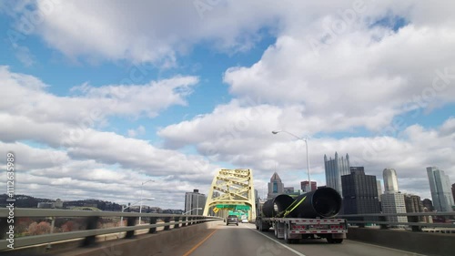 Car driver pov point of view moving toward Pittsburgh, PA city skyline and skyscraper buildings on multilane highway and interstate through vehicle traffic on golden bridge arches with blue sky