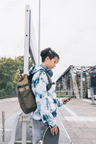 Casual urban portrait of a teen girl carrying a skateboard, wearing headphones, and focused on their smart phone