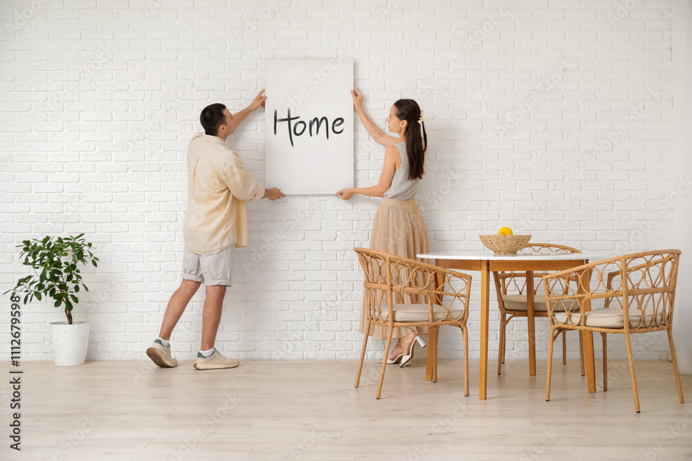 Young couple hanging poster on white brick wall in dining room, back view Young couple hanging poster on white brick wall in dining room, back view