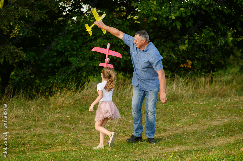 Fototapeta premium Cute girl and middle aged dad having fun outdoors, playing with big toy airplane, running, laughing. Dad and daughter playing tag with airplanes. Childhood , dreams, games. International Father's Day