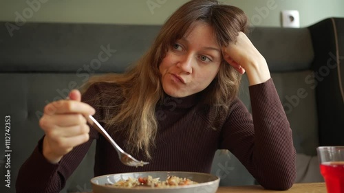 Young woman sits alone at a table, slowly eating her meal. Boredom and unhappiness are evident on her face as she engages in a solitary dining experience