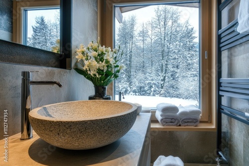 A cozy bathroom featuring a stone sink and floral arrangement with a panoramic view of a serene snow-covered landscape outside a large window.