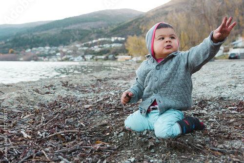 A toddler boy sits on the beach of Lake Wakatipu in Queenstown playing with the mud and the wild ducks while enjoying the outdoors during New Zealand's winter