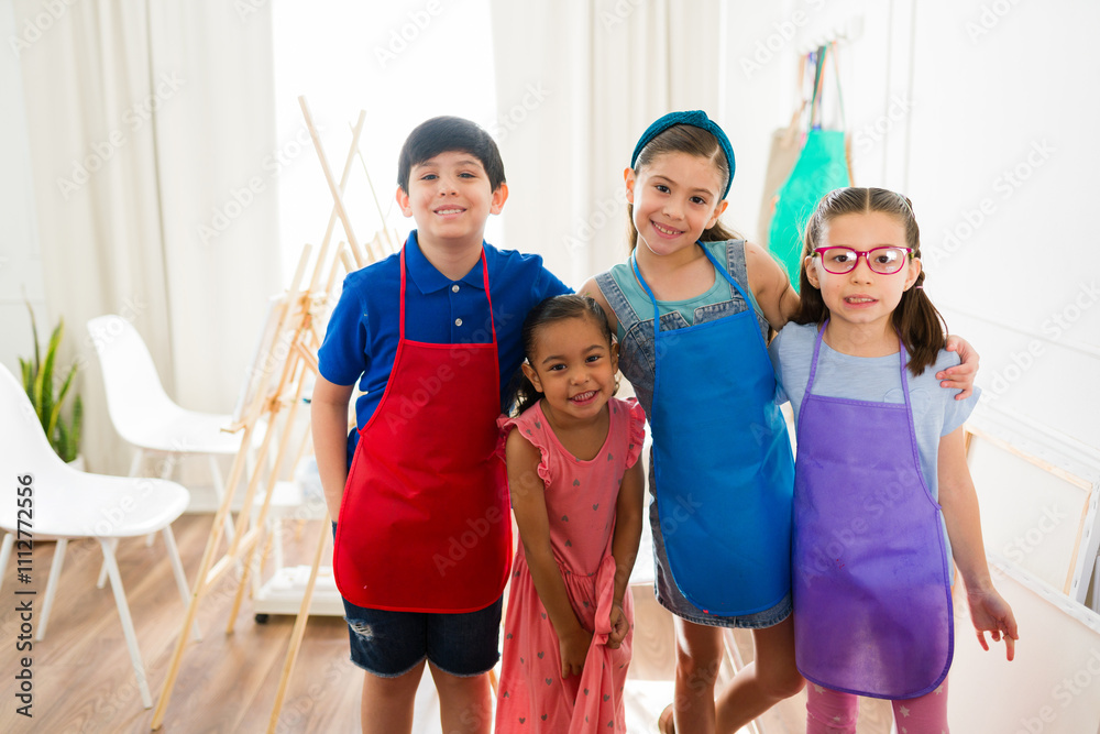 Fototapeta premium Diverse group of happy children wearing colorful smocks standing together in an art classroom, embracing creativity and friendship