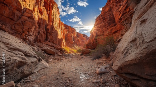 Sunbeams illuminate a narrow canyon, highlighting the vibrant red rock walls and rocky canyon floor.